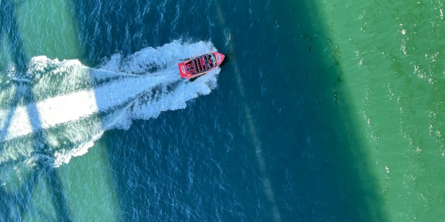 An aerial view of a red jetboat sailing across blue-green water and two long shadows of a bridge overhead in the sunny, Sydney Harbour. 