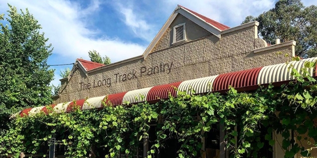 Sandstone building with The Long Track Pantry painted on the street frontage and creeper vines hanging from the red and white striped bullnose veranda