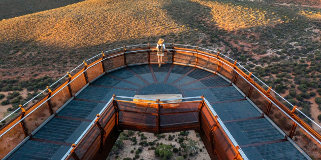 A v-shaped glass and metal skywalk extends over a panorama of scrubby red earth hills at sunset. 