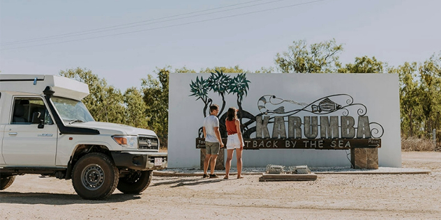  A couple stands on a road next to an SUV and a sign that says Karumba, outback by the sea, framed by trees and blue sky.