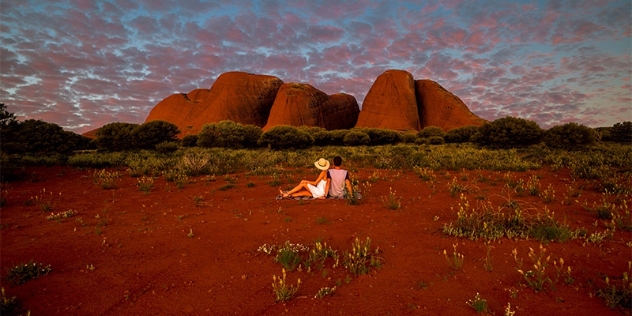 Man and woman sit on the dark ocre desert earth looking at formation of red rocks framed by low green rounded shrubs and with an early evening blue sky with pink clouds