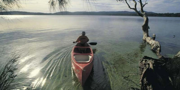A man kayaking near the shore of a still lake surrounded by treed-covered hills.