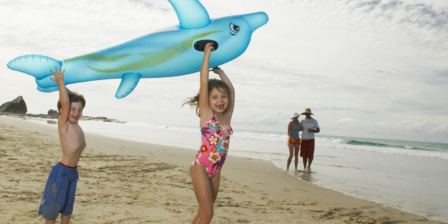  A boy and girl play with an inflatable dolphin toy on a sandy beach, as their parents look on.