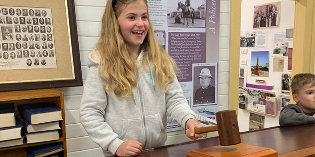 A blond girl banging a wooden gavel on a wood table  in a country town museum with photos on the walls, while a boy stands next to her.