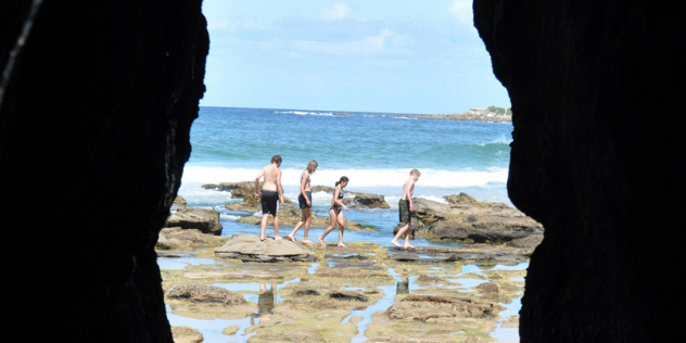  Three boys and a girl walking along rocks on a sunlit beach, as seen through the mouth of a cave. 