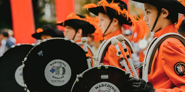 A row of kids in orange uniforms carrying drums that say Marching Koalas, in the Sydney Easter Parade.