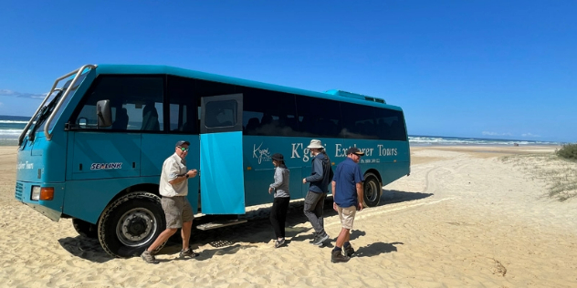  Four people boarding a blue, four wheel drive tour bus on a sunny, sandy beach. 