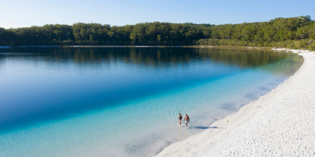 Two people walking along a tree lined, totally white sand beach, leading into clear then deep blue lake waters.