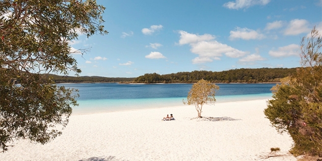 two people sit on a solitary white sandy beach looking out towards clear blue water