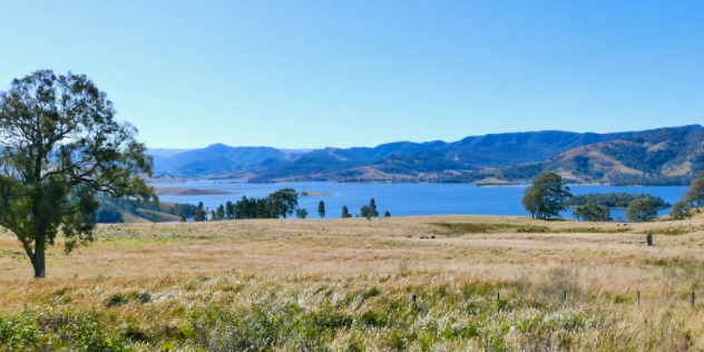  A sunny field dotted with trees leading to an idyllic lake surrounded by tall, green hills. 