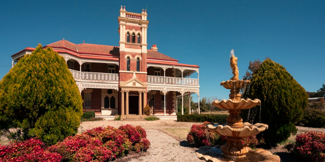 A colonial mansion in coral brick with a wide white verandah, garden and fountain under blue sky.