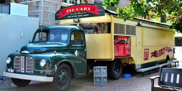 A classic 1940s style green truck with a yellow trailer on the back with red signs and yellow font saying Pie Cart and menus along the side, parked on a sunny city street. 