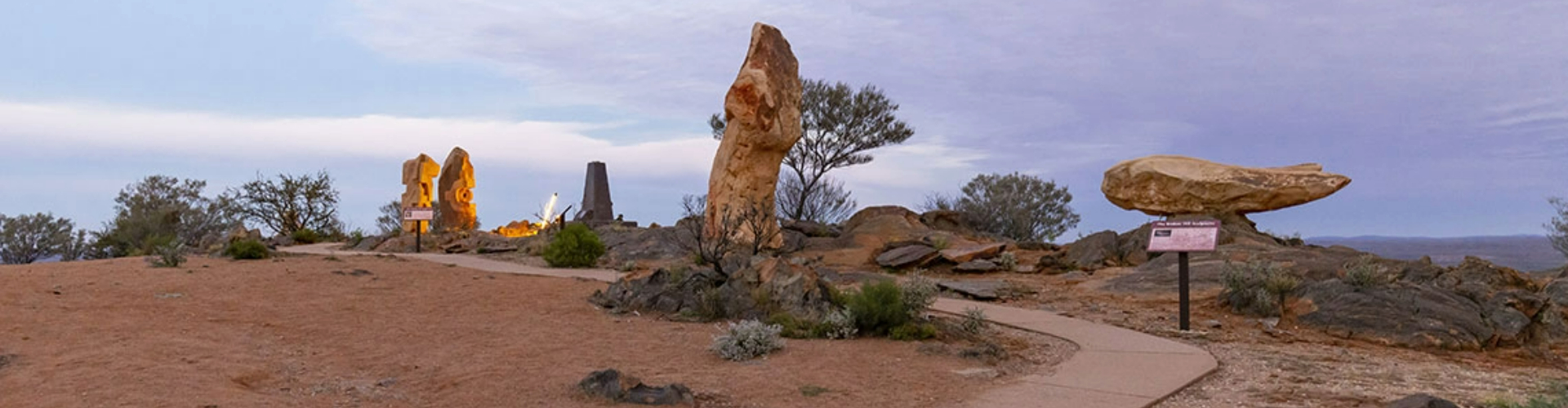 living-desert-sculptures-broken-hill-1920x500 A sidewalk curving up a bare, rocky hill, lined with various roughly carved, pillar-like rock sculptures.