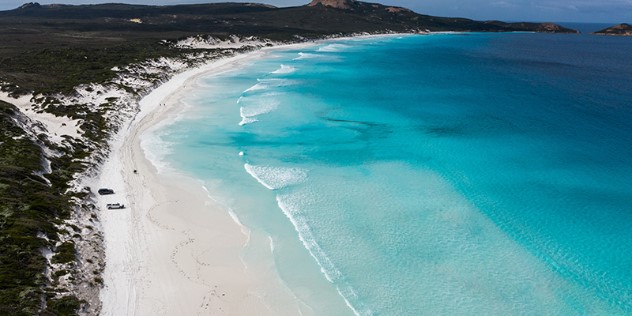 A tree lined, curved white sand beach with two suvs parked near the treeline, surrounded by turquoise waves leading out to dark blue ocean.