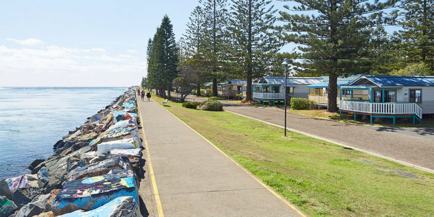  A paved walking path along a rocky shore with art painted on the rocks, next to a row of beach houses with verandas.