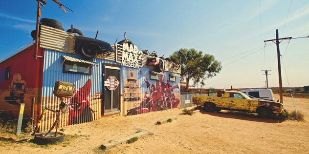 A corrugated-metal building in the dusty outback, painted with in bright-warm colours with scenes from the Mad Max films, with a yellow truck parked out front under a clear, blue sky.  