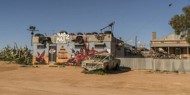  A square corrugated metal building covered with a mural of Mad Max 2 characters, on a sunny dirt road.