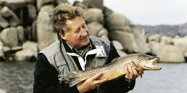 A middle aged blond man holding a trout by a rocky seaside on a cloudy day.