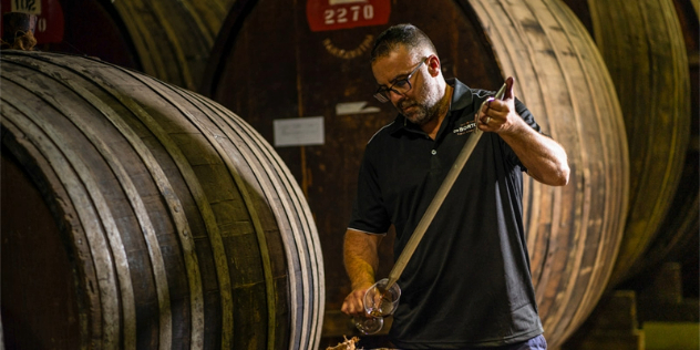 A middle-aged man with a goatee stands in a dim warehouse surrounded by oversize barrels of wine, using a long syringe-like instrument to fill a wine glass. 