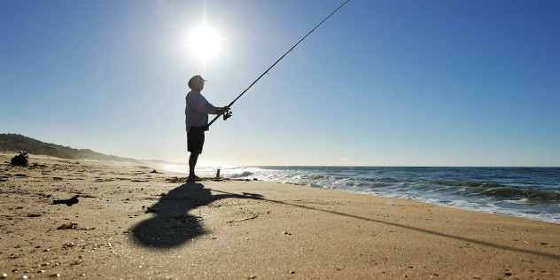 A man silhouetted as he fishes along a sandy shore with the sun blazing down directly above him. 