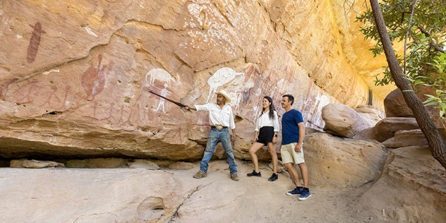 An aboriginal man pointing to a large rockface covered with Indigenous art, as a couple looks on and listens. 