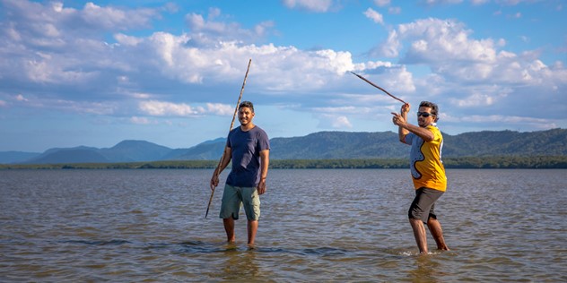 Two men stand in shallow water on a sunny day, both holding spears, one at his side, the other poised to throw the spear. 