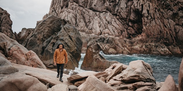 A man with long, dark hair being blown back by wind, wearing an autumn jacket walks along a rocky beach, with rock walls, an inlet of dark blue water and grey skies in the background.