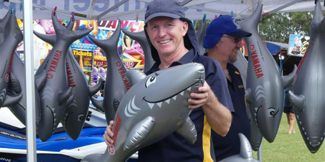  A middle aged man in a cap holds a metre long inflatable shark toy under a market tent full of the same toy.