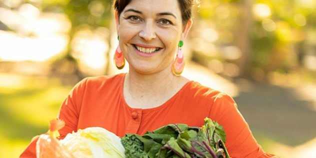 A woman with short, auburn hair smiling and holding a wooden box of groceries.