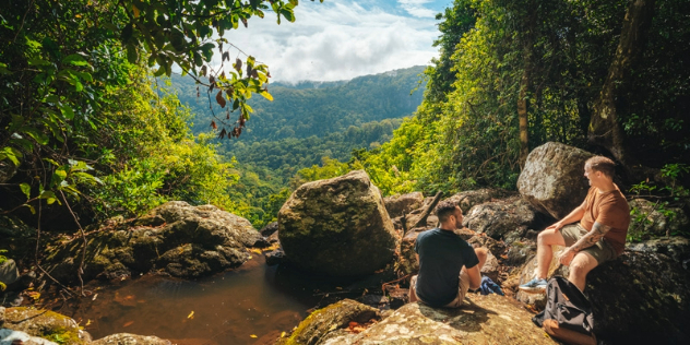 Two men in shorts sit on a cliff surrounded by foliage and small pond looking over forested hills.