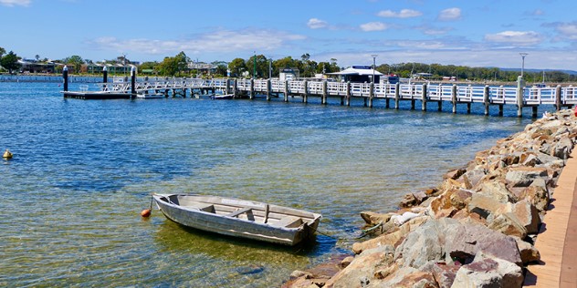 A lone paddleboat floats, tied along the rocky shore near a boardwalk that leads to a jetty, and green trees on the opposite shore.