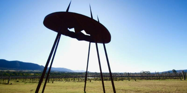  A metal sign supported by metal spikes, with mountain shapes cut out of it, in front of a sunny vineyard. 