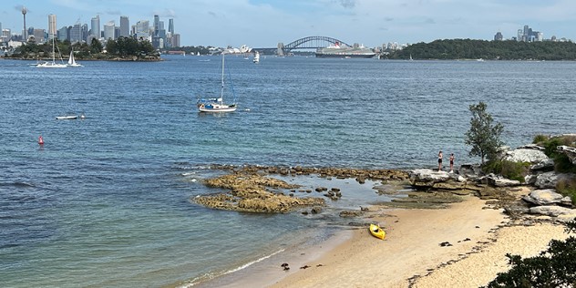 In the foreground is a sandy beach flanked by trees, with an outcrop of rocks that leads out to a broad view of boats on the Sydney harbour, with the city skyline, Harbour bridge and Opera House visible on the horizon.