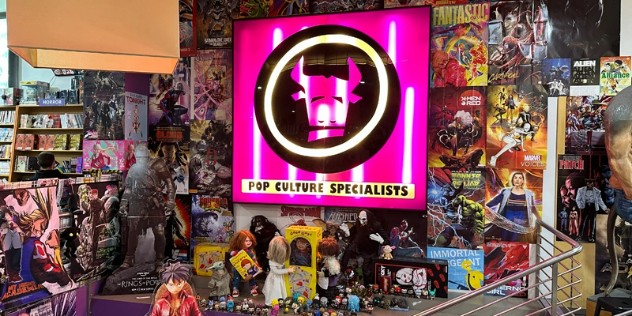 Inside a comic and collectibles store, with a large stylised pink logo of a minotaur's head on the wall. 