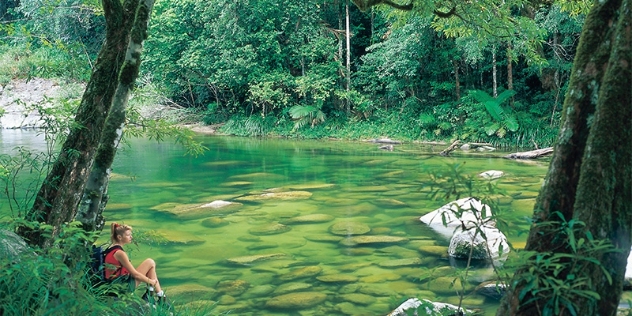 a woman rests against a tree on the river bank and looks out to a shallow green pool surrounded by rainforest 