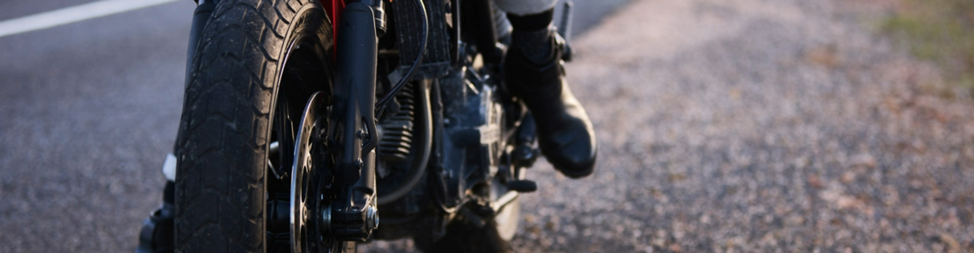 motorbike-on roadside-1920x500 Close up of the front of a motorbike along a roadside with a biker's boot on the kickstand ready to take off.