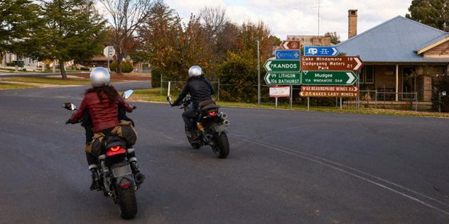 Two motorbike riders in helmets come take a corner near a small town and road signs pointing in all directions.