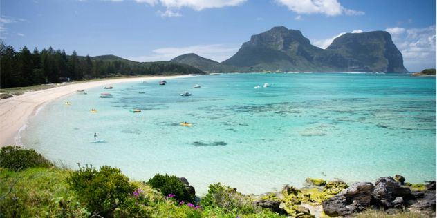 Two treed mountains rising out of turquoise blue ocean into a blue sky in front of a near-circular white sand beach surrounded by lush trees.