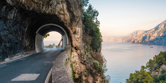 A paved road at the mouth of a short tunnel through a mountainside, to the right is the sea below and more mountains in the distance.