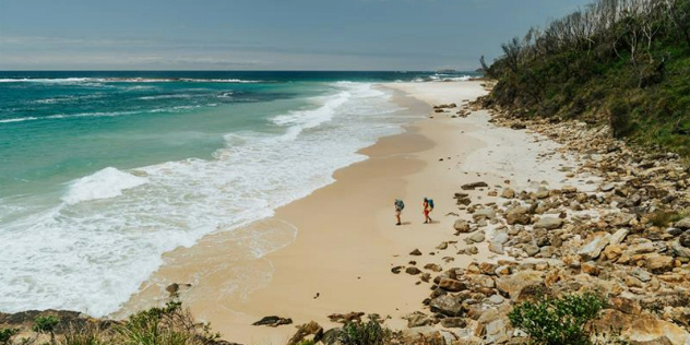  Two hikers appear small as they walk along a wide sand and rock beach with green hills rising from the shore.