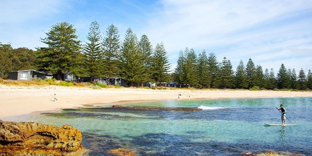 paddleboarder paddles towards the beach on clear water with NRMA holiday cabins in the background