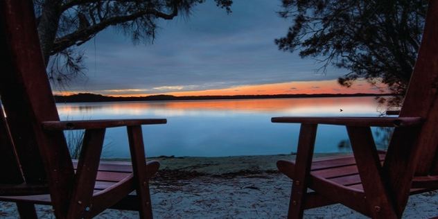 two chairs on the shoreline look out to a sunset on the lake