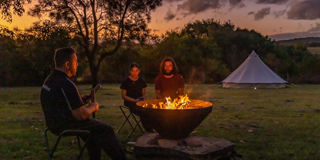 An Aboriginal guide speaks to a couple across a crackling firepit during an orange sunset at a campsite in Gulaga, New South Wales.