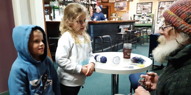 two small children listen intently to a local man as he recalls a Banjo Paterson poem while weaving a scarf inside the Federal Hotel