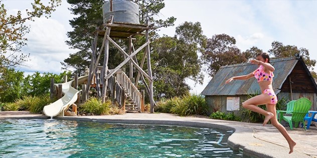  A girl jumps into a pool next to a waterslide wrapped around a miniature water tower, framed by trees.