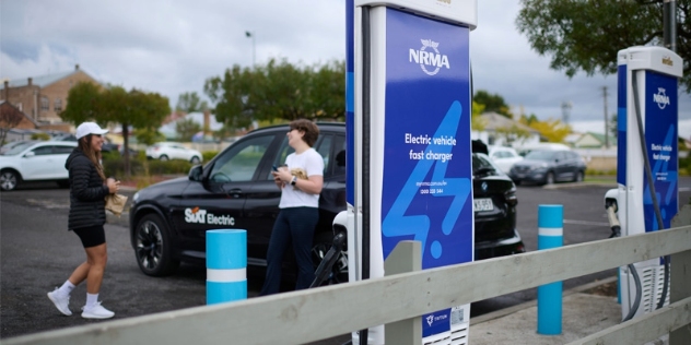 black electric vehicle with SiXT logo on the side plugged into an NRMA fast charger with two people holding snacks and talking while they wait