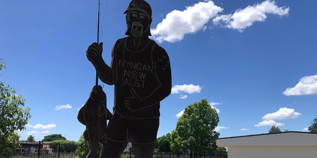 large metal cut out of a man with beard, cap and mullet hair holding a fish and wearing a vest that says Nyngan NSW Aust towers above a nearby building