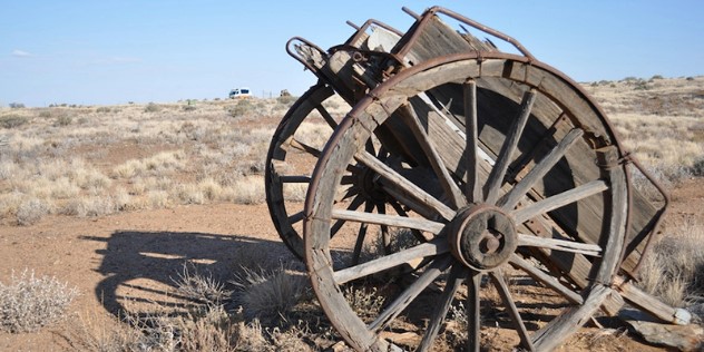 An old, wooden farming wagon with large wheels sits tipped forward on a near-barren brown earth hill with patches of scrub, with a combi van in the distance and blue sky on the horizon.