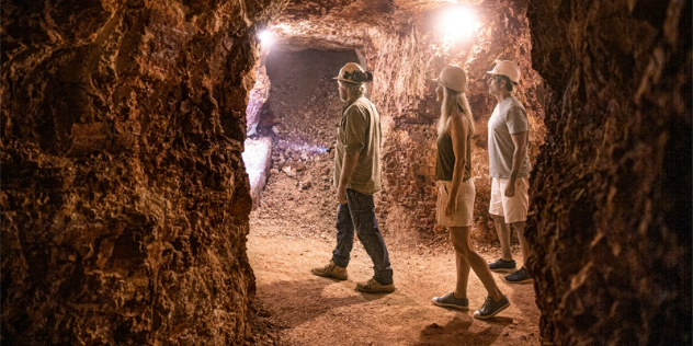 Two men and a woman in hard hats walking upright through a rocky mine tunnel that is lit by warm white lights screwed along the tunnel walls.