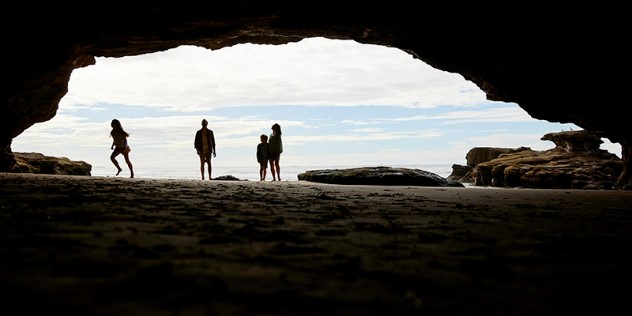 The curved, but rocky open mouth of a cave as seen from inside, a family parents and children are silhouetted along the entrance, with cloudy blue sky in the background. 